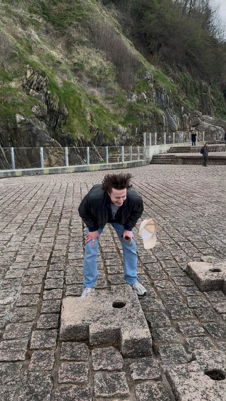 Mo standing above a tube that is blowing air in his face and his hat falling down beside him